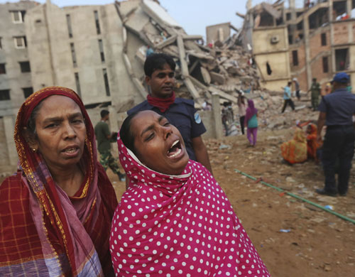 Two women grieving with collapsed factory in background.