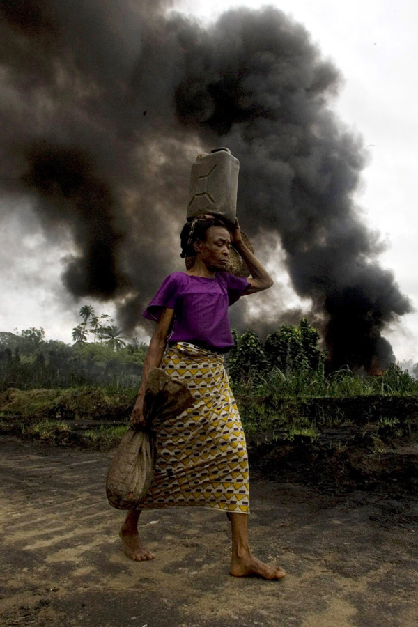 A woman walks past a burning oil pipeline in Kegbara Dere, Nigeria, in 2007. The fire burned for 45 days and 45 nights, blanketing the village with ash and torching the young cassava plants.
