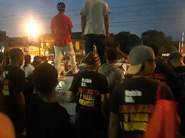 People stand on the hood of a police car surrounded by others protesting against police murder.