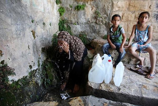 Older Palestinian woman waits with two children to fill water bottles during water crisis.