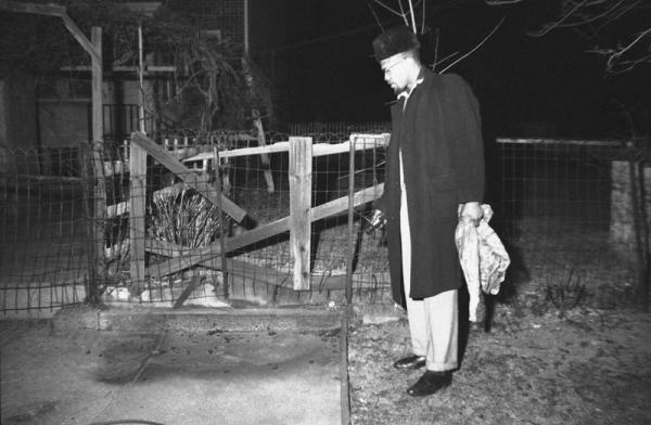 Malcolm X examines damage to sidewalk in front of his home from molotov cocktail. 
