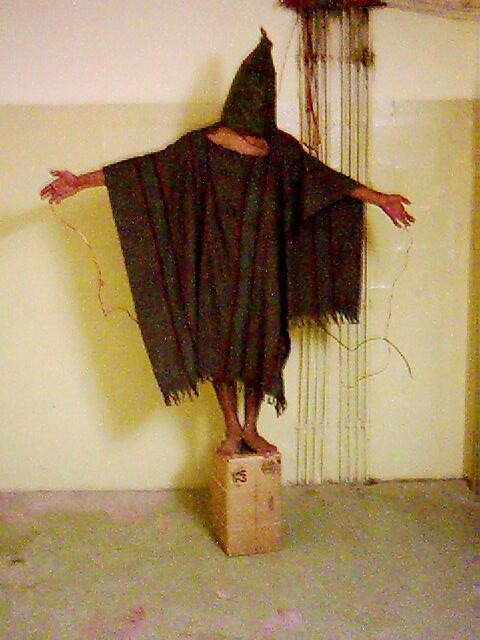 Torture at Abu Ghraib: a hooded Iraqi prisoner, forced to balance on a small box, with wires attached to his fingers—he was forced to stand for hours, told that if he fell over, the wires would electrocute him.