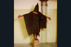 , a hooded Iraqi is shown balanced on small box, with wires attached to his fingers—he was forced to stand for hours, told that if he fell over from exhaustion, the wires would electrocute him